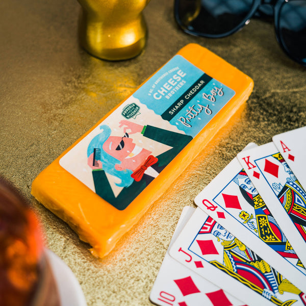 A packaged block of 'Pretty Boy' sharp cheddar cheese with a blue label, placed on a glamorous setting with a deck of cards and sunglasses in the background.