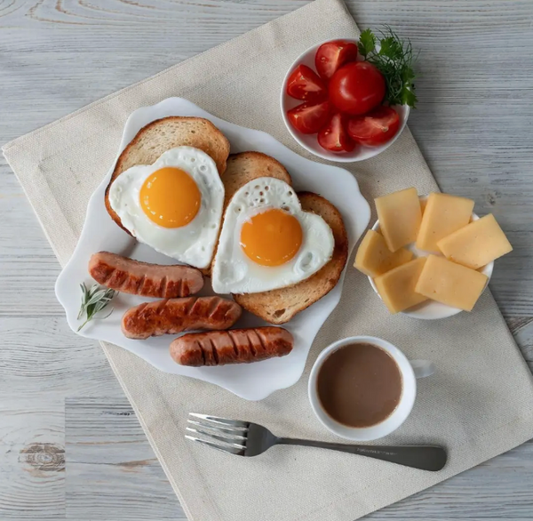Breakfast setting with heart-shaped eggs, sausages, cheese, and coffee on a wooden table.